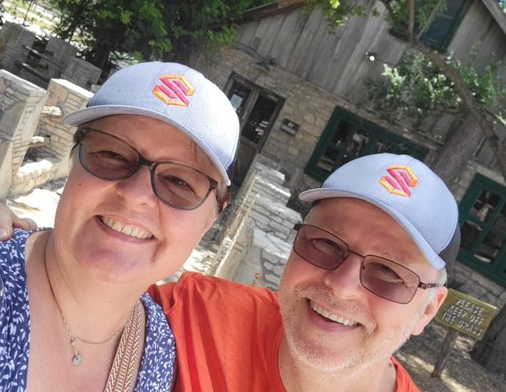 Two people smiling outdoors, both wearing glasses and grey hats with an orange logo, standing in front of a stone building and trees.