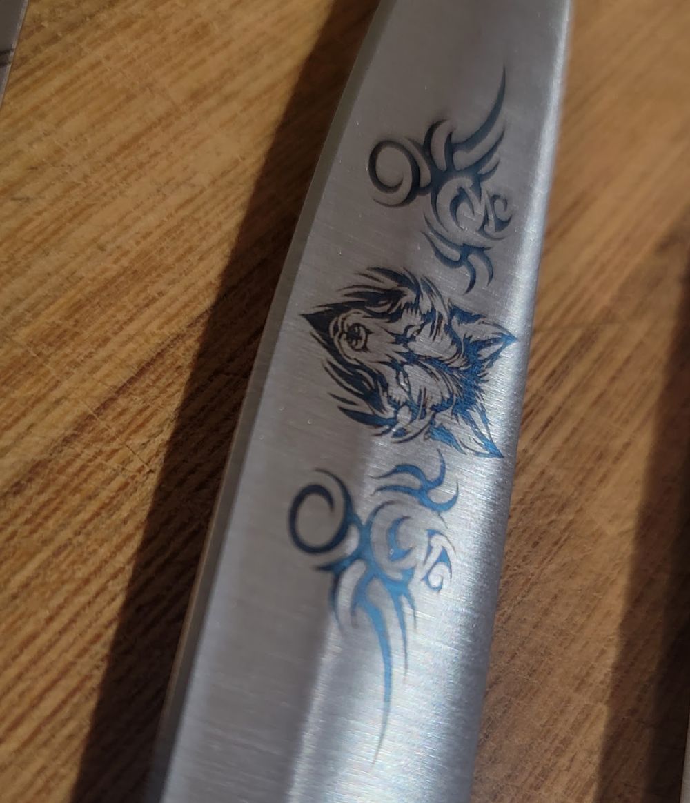 A close-up of a knife blade with a detailed wolf and tribal design etched into the metal, resting on a wooden surface.