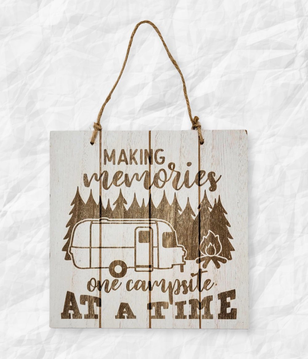 A wooden sign with an illustration of a camper, trees, and a campfire, displaying the text “Making memories one campsite at a time.”.