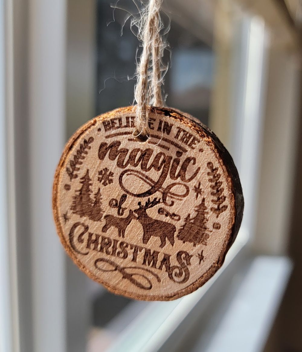 A round wooden ornament with the text "Believe in the magic of Christmas," hanging by twine, featuring engraved reindeer, trees, and snowflakes.