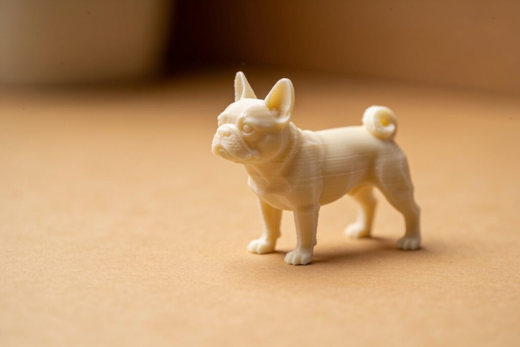 A small, beige 3D-printed figurine of a dog resembling a French bulldog stands on a light brown surface.